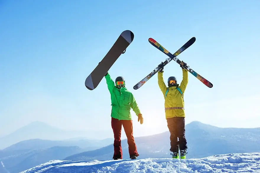 Snowboarder and skier holding their snow equipment above their heads in triumphant on top of the Snowy mountains.
